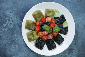 White plate with black and spinach ravioli, cherry tomatoes and green basil leaves, flatlay on a blue stone surface, horizontal shot