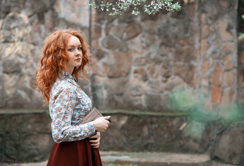 young red-haired curly-haired girl with freckles posing in a summer garden with a vintage book in her hands