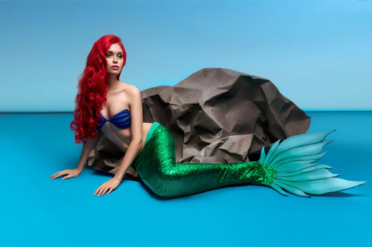 Mermaid with red hair resting near stone on blue background