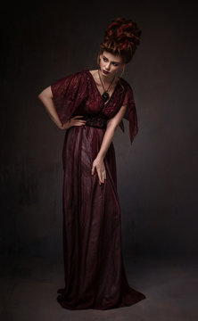 Full length view of woman with baroque hairstyle and evening maroon dress posing on dark background