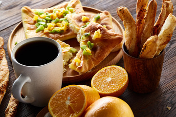 Morning concept with a cup of coffee, breadsticks, fruit pies and oranges on wooden background.