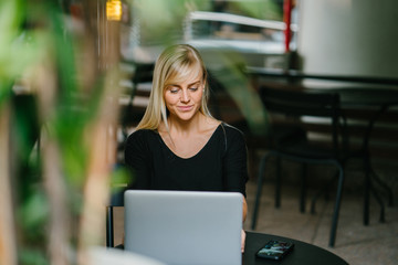 A young and attractive blonde woman is working on her laptop inside a cafe. She is sitting down and smiling as she concentrates on her task.