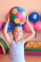 A little girl in the gym is holding a big ball.