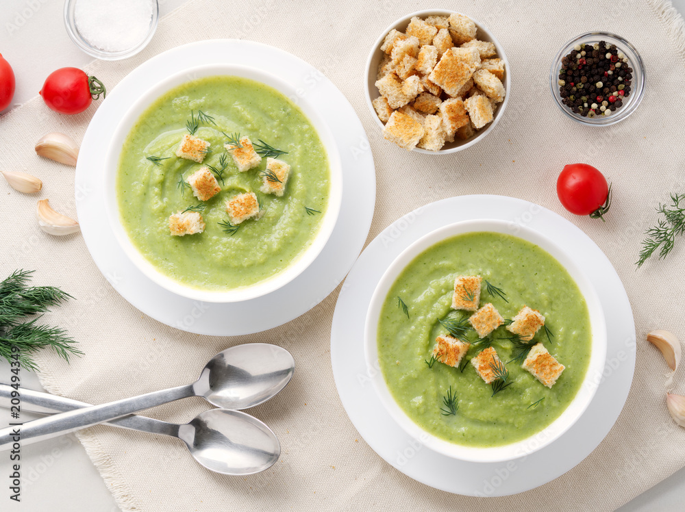 Wall mural Two large white bowl with vegetable green cream soup of broccoli, zucchini, green peas on white background, top view