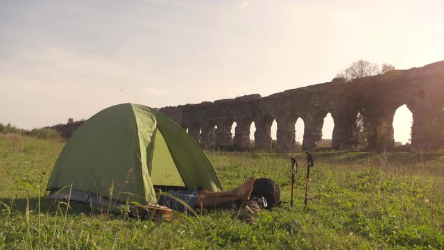Young man backpacker sleeping in camping tent on grass with feet outside in front of roman aqueduct arches in parco degli acquedotti park ruins in rome at sunset sticks in ground slow motion