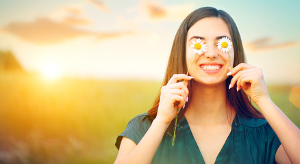 Beauty joyful girl with daisy flowers on her eyes enjoying nature and laughing on summer field. Beautiful young woman having fun