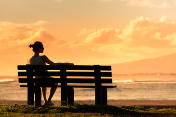 Thoughtful young woman relaxing on park bench watching the ocean sunset. 