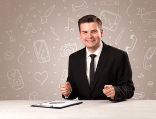 Young handsome businessman sitting at a desk with white mixed media icons behind him