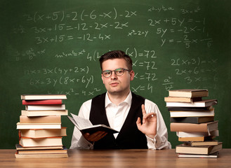 A young ambitious teacher in glasses sitting at classroom desk with pile of books in front of blackboard full of math calculations, numbers, back to school concept.