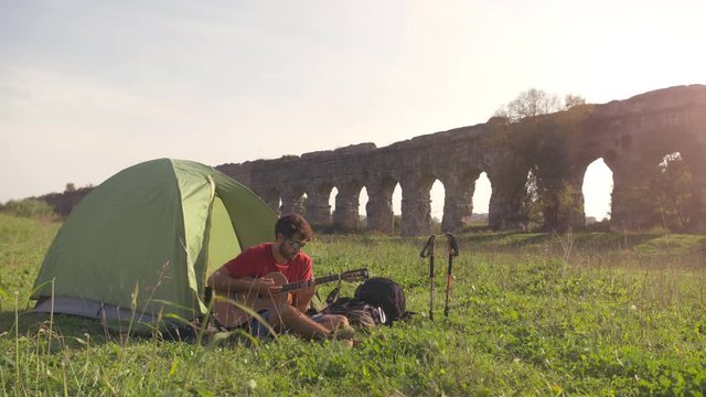 Young man backpacker playing guitar outside camping tent sitting on grass in front of roman aqueduct arches in parco degli acquedotti park ruins in rome at sunset sticks in ground slow motion