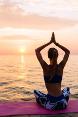 Fototapeta na wymiar Yoga at sunset. Young woman in sportswear, is engaged in yoga, sitting on pier in lotus pose, meditating, looking at sea sunset. Relaxation time