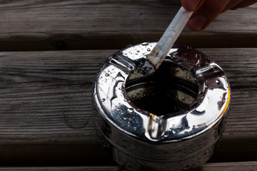 Burning cigarette in a metal ashtray. Healthcare concept with smelly and dirty cigarette. Lung cancer warning for smokers. Hand holds burning cigarette above ashtrey.