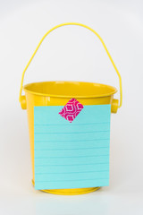 Aqua colored lined paper list taped to front of bright yellow miniature bucket with solid white background to illustrate bucket list