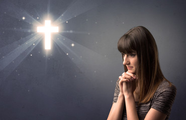 Young woman praying on a grey background with a shiny cross above her