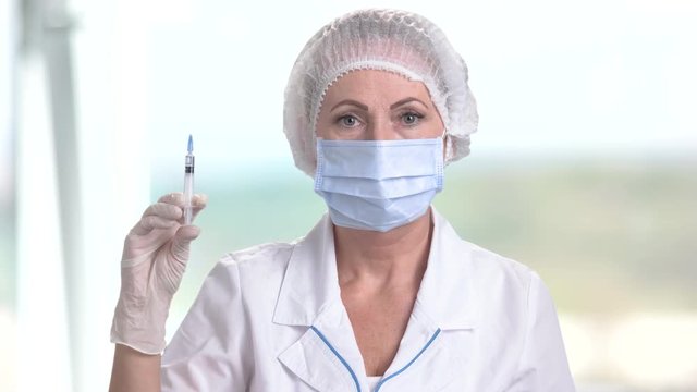 Nurse or doctor holding medical syringe. Portrait of female doctor wearing surgical cap and face mask standing with injection on blurred background.