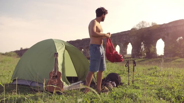 Young man backpacker dressing wearing t-shirt outside camping tent sitting on grass in front of roman aqueduct arches in parco degli acquedotti park ruins in rome at sunset sticks in ground slow