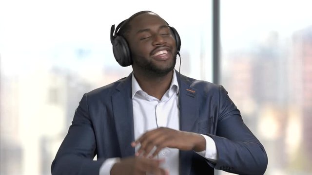 Black manager in headphones is dancing. Joyful dark-skinned businessman listening to music in headphones. Enjoy of music concept.