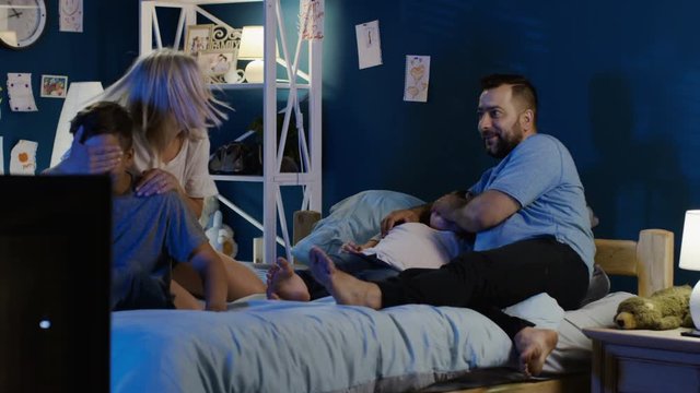Adult couple watching TV with kids and avoiding erotic scene by switching channel while lying together on bed