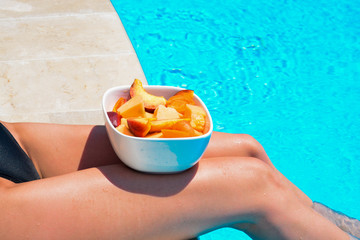 Tanned beautiful legs. Young woman relaxing and eating fruits. Fruits in a bowl by the pool near a thin slender fit girl legs. Fruit salad, healthy food. Summer concept.