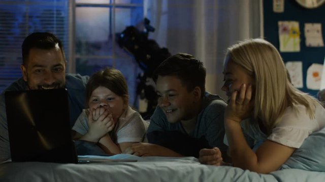 Excited parents with children chilling on bed at home in evening time and watching comic show on laptop all together