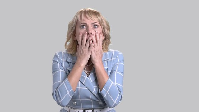 Shocked horrified woman on grey background. Portrait of terrified mature female holding hands on head. Human expression of horror.