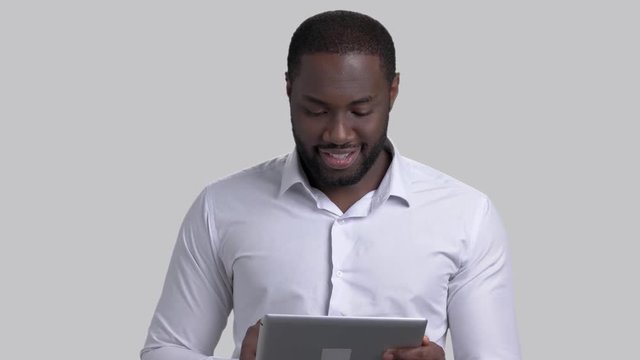 Cheerful afro-american businessman using electronic tablet. Black smiling man in white shirt with computer tablet on grey background.