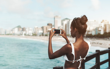 View from behind of an exquisite Brazilian girl in swimsuit using her cellphone to take panoramic photo of coastline; rear view of a young svelte black female with a smartphone photographing the beach - obrazy, fototapety, plakaty