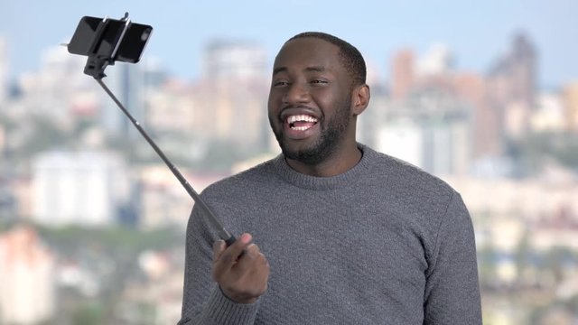 Funny african-american guy using selfie stick. Young attractive dark-skinnes man taking selfie with monopod on blurred background.