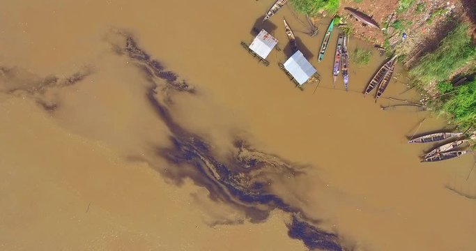 Aerial Overhead Of An Layer Oil Or Petrol From An Engine Boat Across The Water Surface.  Houses Boats And Dugout Canoe At The Edge The River  