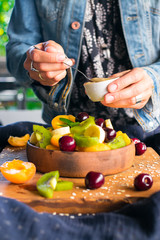 Woman hands adds sweet banana smooth cream with spoon to fruit salad in bowl on wooden board. Healthy sweet dessert for breakfast. Raw vegan vegetarian healthy food