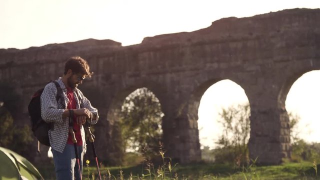 Young man backpacker walks barefeet with sticks on grass in front of roman aqueduct arches in parco degli acquedotti park ruins in rome at sunset slow motion