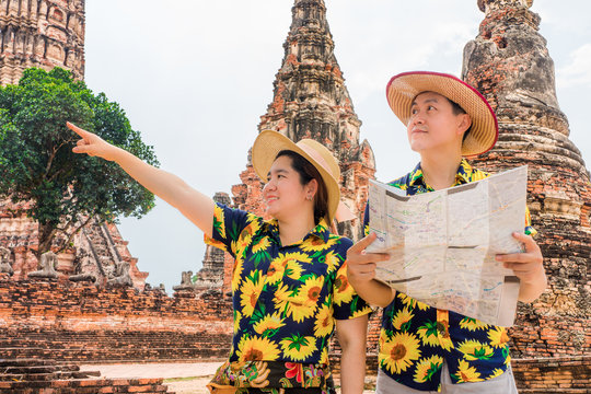 Asian tourists with colorful floral shirts using paper map to find attraction in old temple ruins. Couple go sightseeing in Wat Chaiwatthanaram, Ayutthaya Historical Park, Thailand, Southeast Asia.