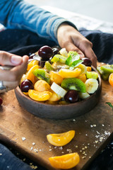 Woman hands holds fruit salad in bowl on wooden board. Healthy sweet dessert for breakfast with rip banana, apricot slices, fresh kiwi, cherry, mint leaves, cream. Raw vegan vegetarian healthy food