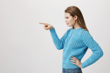 Indoor portrait of beautiful blonde girl standing in profile with hand on waist, pointing left and looking at that direction, over gray background. Woman indicates on high heels she desires to buy