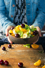 Woman hands holds fruit salad in bowl on wooden board. Healthy sweet dessert for breakfast with rip banana, apricot slices, fresh kiwi, cherry, mint leaves, cream. Raw vegan vegetarian healthy food