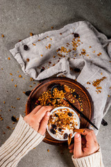 Ideas for an autumn winter breakfast. Spicy yoghurt with granola, dried berries, nuts, almonds, spices (cinnamon, anise), in cup, on plate. Gray background, girl hold mug in hands. Top view copy space
