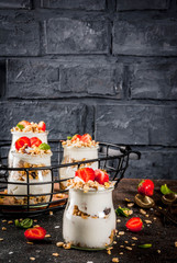 Healthy summer breakfast idea, homemade layered parfe dessert in small jar with granola, yoghurt and strawberry, dark background copy space