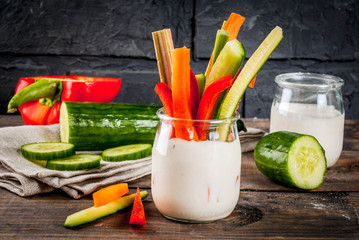 Summer healthy snack appetizer, assorted colorful fresh vegetable sticks (celery, rhubarb, pepper, cucumber and carrot) with yoghurt sauce dip, wood and dark concrete background copy space
