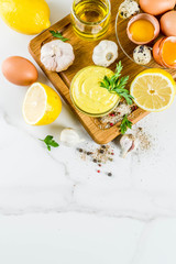 Homemade mayonnaise sauce with ingredients - lemon, eggs, olive oil, spices and herbs, white marble kitchen table copy space