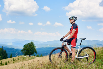 Naklejka na ściany i meble Athletic sportsman biker standing with cross country bicycle on grassy valley on summer day, enjoying beautiful view of Carpathian mountains on background. Healthy lifestyle and outdoor sport concept