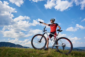 Fototapeta na wymiar Professional male cyclist in sportswear and helmet standing with cross country bike on top of hill, pointing to the blue sky with clouds on summer sunny day. Active lifestyle and outdoor sport concept