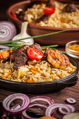 The concept of eastern, Uzbek cuisine. Pilaf, plov in a black cast-iron frying pan on a wooden table, next to lie ingredients, onions, tomatoes, parsley, pepper. Central Asian cuisine.