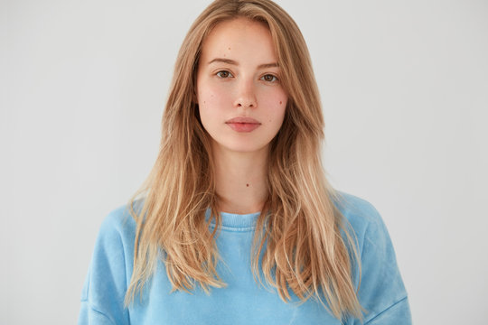 Portrait of beautiful young female with pleasant appearance looks seriously at camera, has pure skin, listens information from interlocutor, wears blue sweater, isolated over white background.