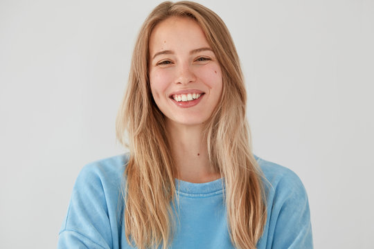 Portrait of happy lovely female has broad smile, being in good mood after party with friends, has long hair, wears casual oversized sweater, isolated over white background. Positive emotions concept
