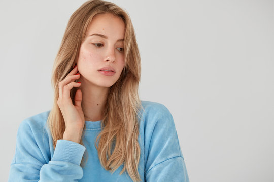 Thoughtful light haired female looks pensively aside, focused down, wears blue sweater, thinks about something, contemplates about future plans, stands against white background with blank space
