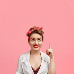 Photo of attractive cheerful female with charming smile, has red lips and white teeth, indicates upwards with fore finger, looks cheerfully on ceiling, isolated over pink background. Style concept