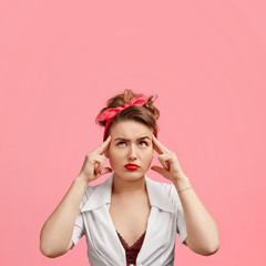 Concentrated beautiful young female keeps fingers on temples, looks thoughtfully upwards, tries to concentrate on something, poses against pink background. People, thoughts and lifestyle concept