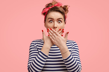 Beautiful surprised female with make up, holds palms on lips, looks with eyes popped out, wears striped sweater and headband, expresses great shock while recieves unexpected news, isolated on pink