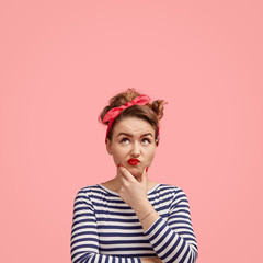 Vertical shot of pretty female with confused expression, curves lips and looks thoughtfully upwards, poses against pink background with copy space, holds chin and curves lips has make up, red manicure