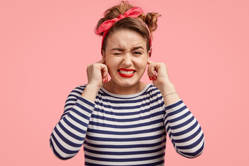 Omg, what sound! Stressful beautiful female plugs ears and frowns face, hears something annoying, wears sailor striped jacket, isolated over pink background. People, negative feeling concept